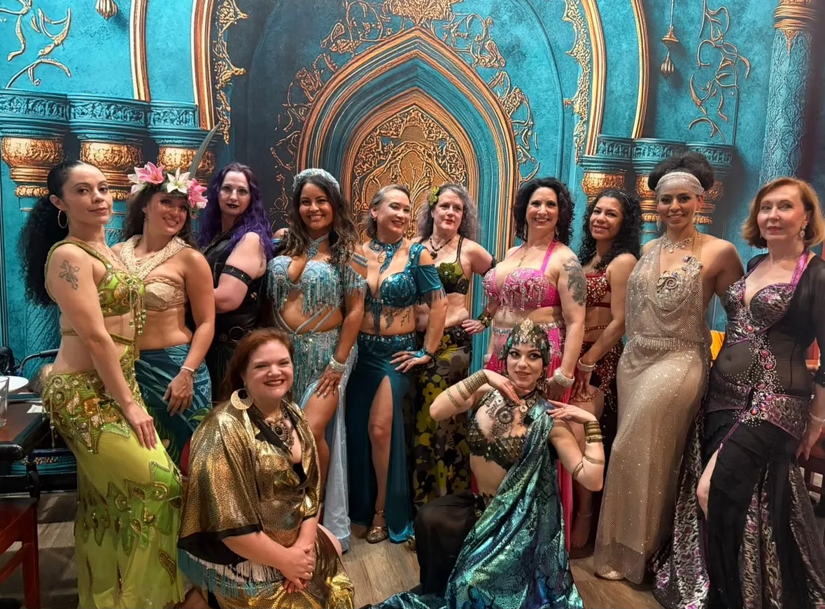 A group of belly dancers in bright sequined costumes posing together in front of the mural at Marrakesh DC