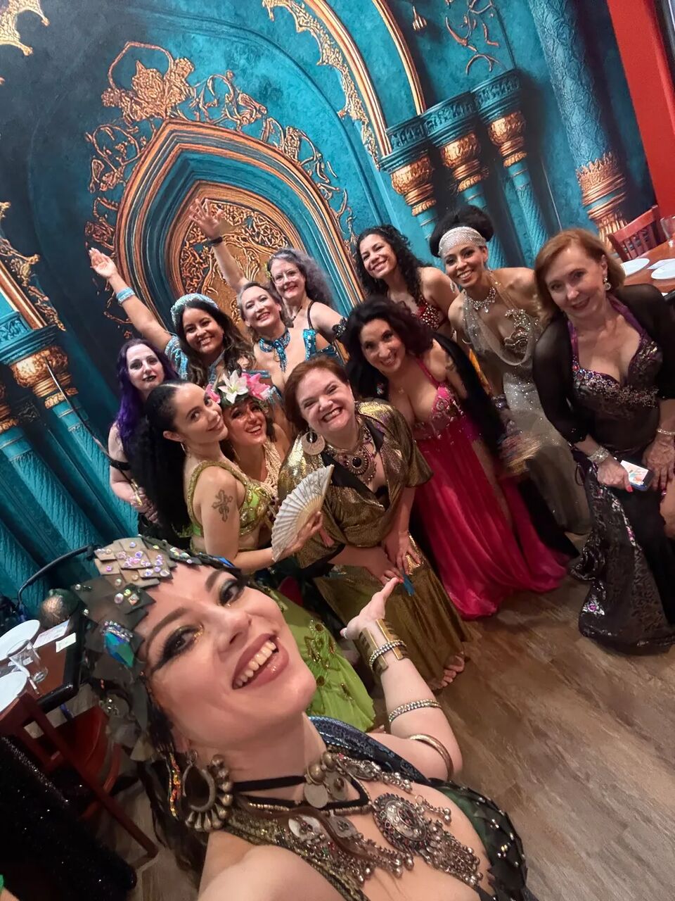 Group selfie of dancers in sparkling costumes by the teal archway backdrop at the August Hafla