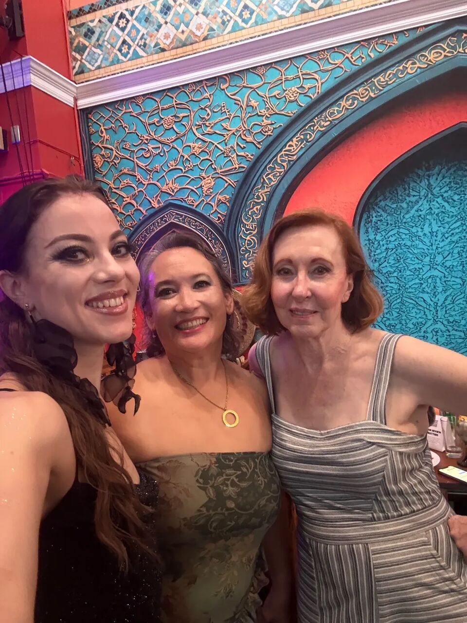 Three friends smiling for a selfie in front of the ornate teal and red wall at the August Hafla