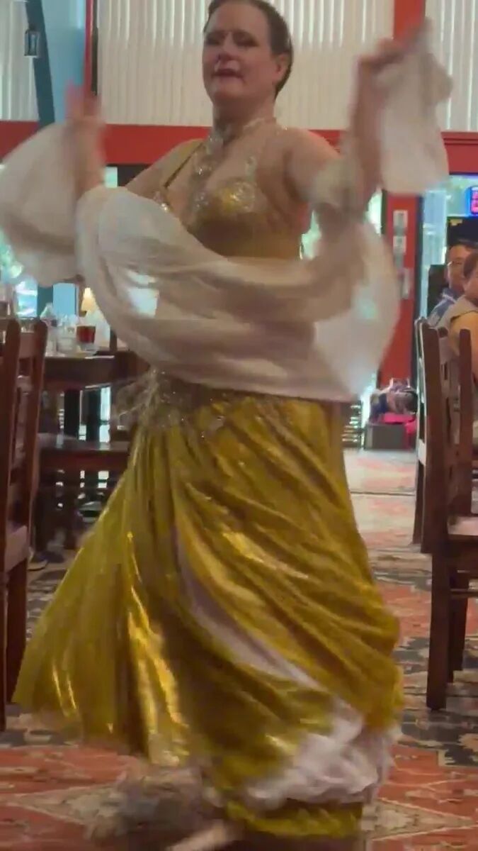 Dancer in a gold costume with white veil sleeves mid-performance at the July Hafla