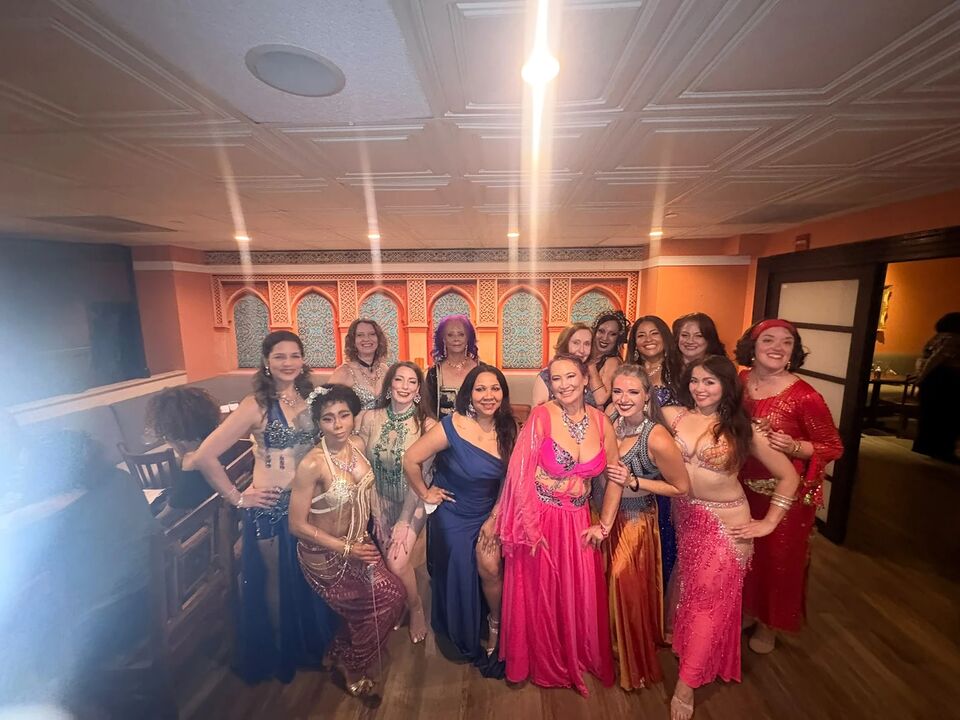 Fifteen dancers in blue, pink, and gold costumes lined up together in the dining room at the September Hafla