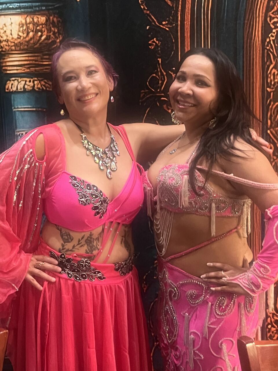 Two dancers in pink and magenta costumes smiling arm in arm in front of the carved wooden screen at the September Hafla