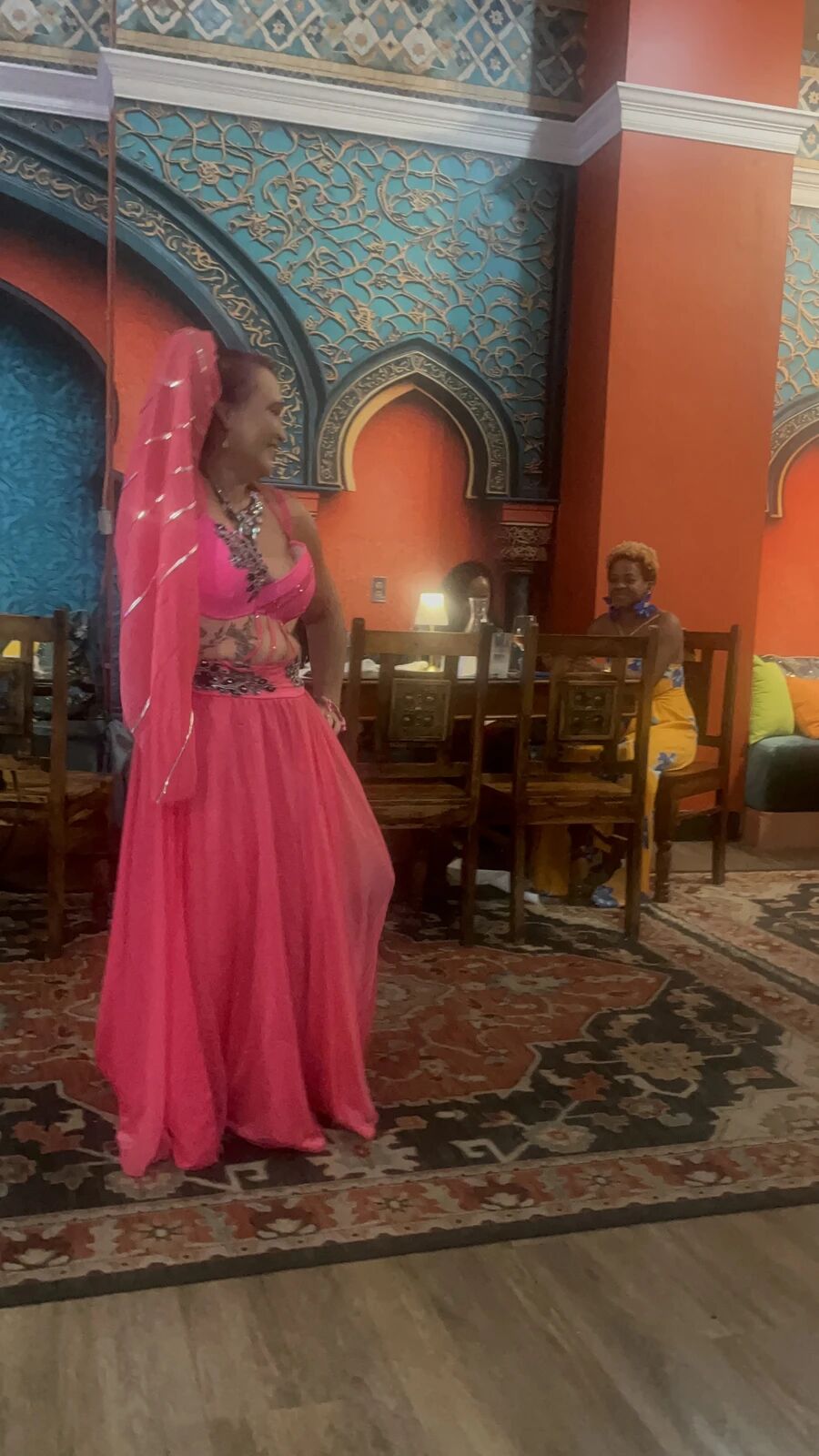 Dancer in a pink costume with a sheer pink veil posing on the rug before the blue arch at the September Hafla