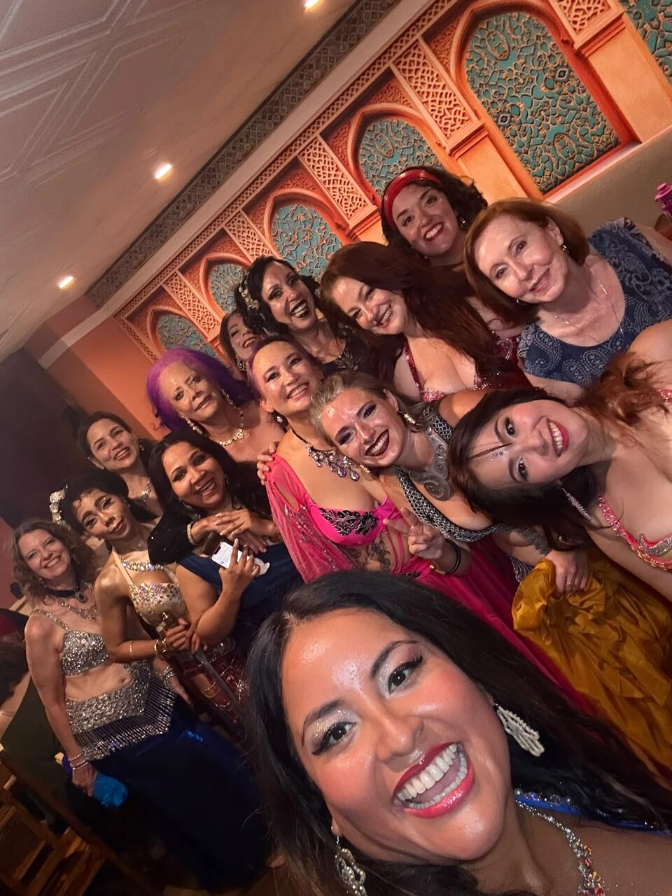 Group selfie of the September Hafla dancers crowded together and laughing beneath the carved arches
